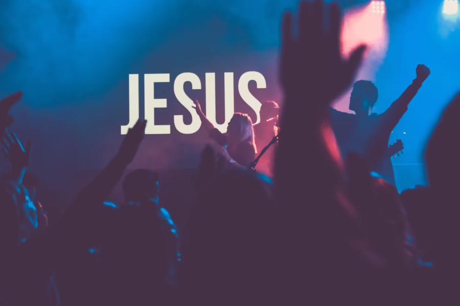 people worshipping with hands in the air. Neon sign reading Jesus